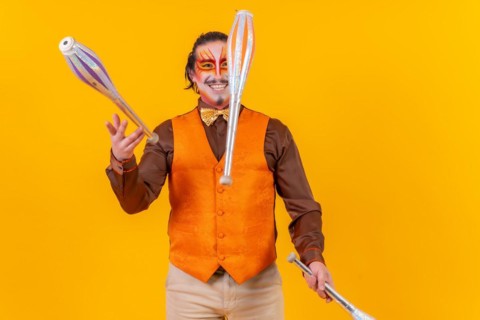 Happy juggler man in makeup vest juggling maces on a yellow background