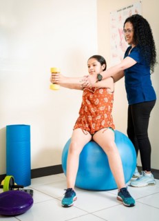 Physiotherapist with female patient holding hand dumbbell sitting on rehab ball. Physiotherapist helping female patient with hand weight sitting on fitness ball