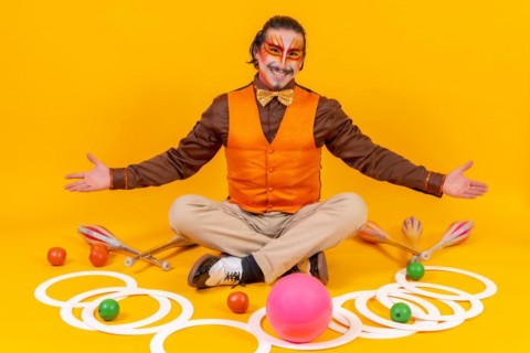Juggler in a vest and with his face painted sitting with the juggling objects on a yellow background