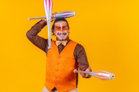 Juggler man in make up vest juggling with maces on a yellow background