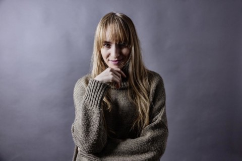 Woman with long blond hair and woollen jumper holds a hand to her chin and smiles at camera