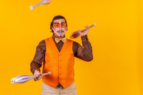 Happy juggler man in makeup vest juggling maces on a yellow background