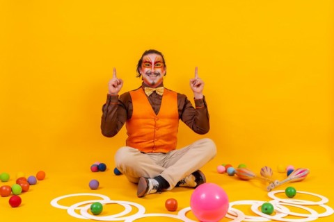 Juggler in waistcoat and with painted face sitting with the juggling objects on a yellow background
