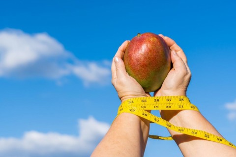 Woman holding a mango in her hands with a tape measure and a sky with clouds in the background