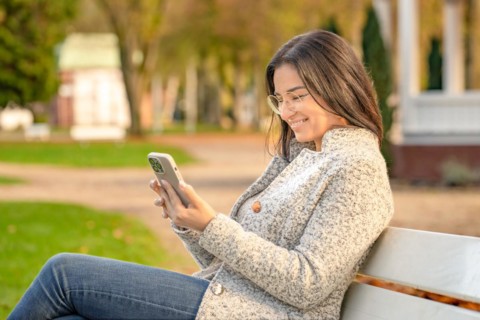 Pretty woman with glasses and Iphone in hand sits on bench in park