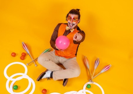 Portrait of a juggler in a vest and makeup sitting with the juggling objects on a yellow background
