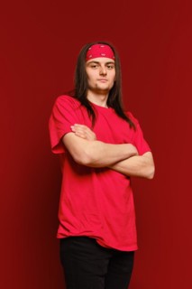 A young adult man in red shirt and head band stands in a studio with arms crossed and looks confidently at the camera against red background