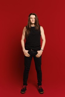Full body indoor portrait of positive young man with long hair standing against red background