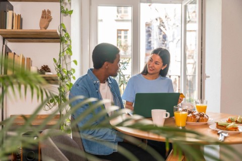 Couple making purchase online with the computer while having breakfast, next to the window, buying flights