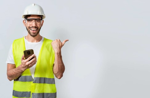 Smiling engineer holding phone and pointing to the side isolated. Young construction engineer holding phone and pointing at blank space. Civil engineer holding cellphone and pointing a promo