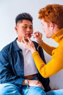 Beautiful gay couple putting on makeup painting their lips being romantic indoors at home, lgbt concept