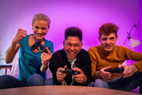 Group of young friends playing video games together on the sofa at home, competing looking at camera