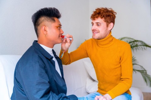 Beautiful gay couple putting on makeup painting their lips, being romantic indoors at home, lgbt concept