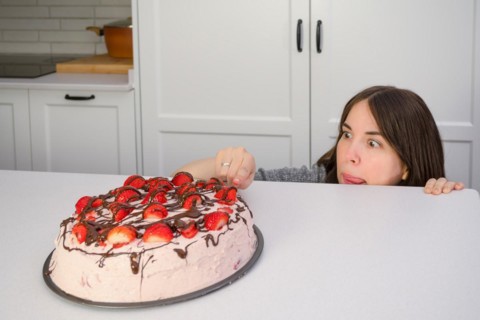 Young woman hiding behind the table eating strawberry shortcake in her home kitchen