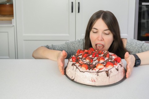 Young woman hiding behind the table eating strawberry shortcake in her home kitchen