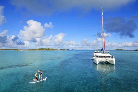 Women paddle SUP and do yoga in front of a sailing catamaran in the harbour of Warderick Wells