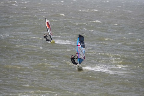 Two recreational windsurfers in black wetsuits practising classic windsurfing along the North Sea coast in windy weather during winter storm