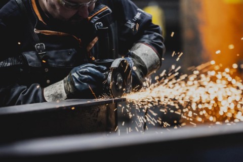 A metal worker works on a steel beam with a cut-off grinder