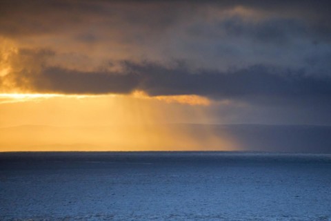 Evening sun breaks through low-lying rain clouds over the Summer Isles and the open waters of the blue Atlantic