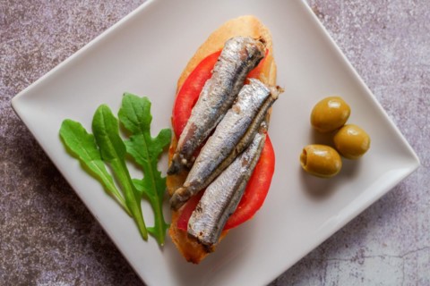 Tapa of sardines on a slice of bread with tomato and olives on a white plate with a typical spanish white background