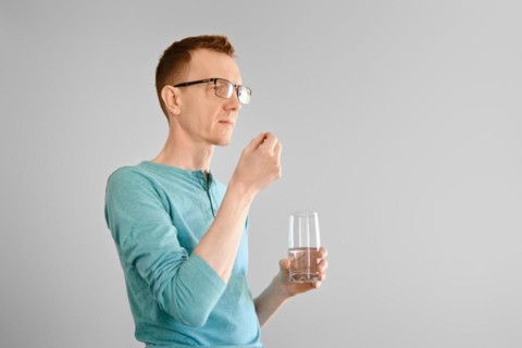 Middle aged man standing with a glass of water and takes pill from headache