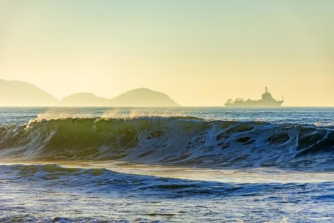 Wave breaking on the beach during sunrise with ship crossing the horizon and mountains in the background.