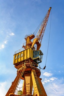 Old and obsolete yellow crane on the harbor pier seen from below and lit by the afternoon sun with blue sky in the background.