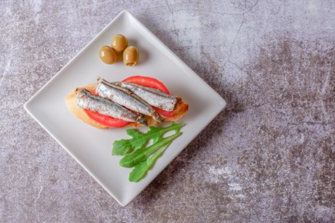 Tapa of sardines on a slice of bread with tomato and olives on a white plate with a typical spanish white background