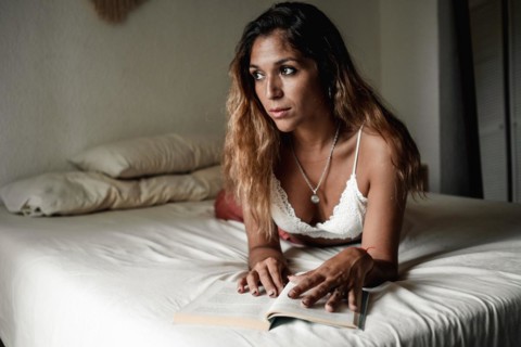 Young latina girl with long hair reading a book lying on her bed is distracted looking towards the window