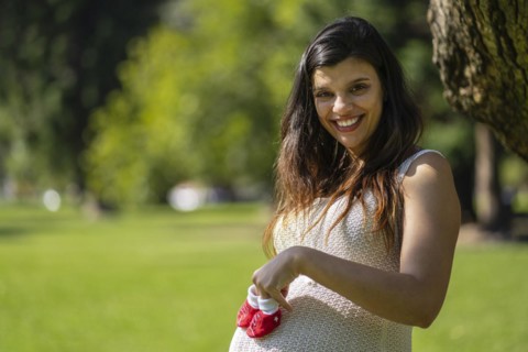 Pregnant latin woman holding baby shoes on her tummy in a park