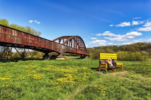 Visitors sitting on large yellow wooden bench in front of old railway bridge