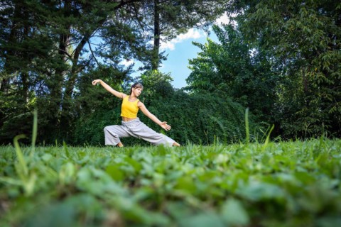 Woman doing qi gong tai chi exercise. A woman practices tai chi in nature surrounded by trees and a blue sky. Close-up of the grass