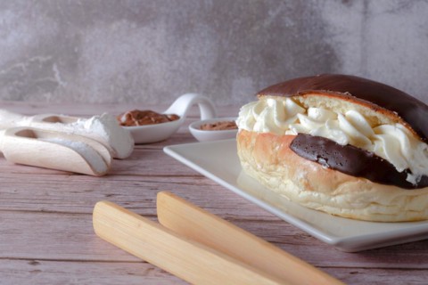 Delicious chocolate-covered cream eclair and white ceramic spoons with chocolate and cocoa on the side with wooden tongs