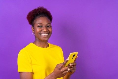 Young african american woman isolated on a purple background smiling with the mobile phone