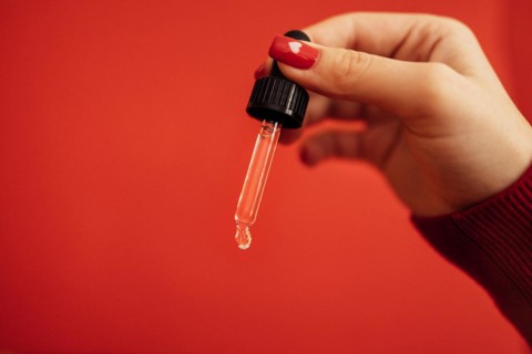 Woman with red manicure holding transparent serum or oil glass pipette. Bright red background. Mock up. Concept of cosmetic and selfcare. Copy space