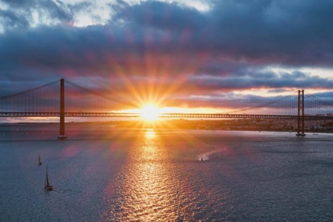 View of 25 de Abril Bridge famous tourist landmark of Lisbon connecting Lisboa and Almada on Setubal Peninsula over Tagus river with tourist yacht boats on sunset. Lisbon