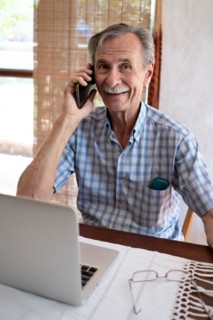 Mature business man smiling and talking on phone in front of laptop looking towards camera