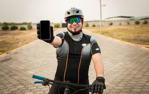 Cyclist in sportswear showing the phone screen. Male cyclist on the bike showing cell phone screen outdoors. Smiling cyclist showing an advertisement on cell phone