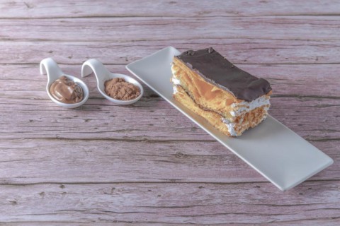 Close-up of a mille-feuille filled with cream and almonds covered with chocolate on a white plate with ceramic spoons with chocolate and cocoa powder