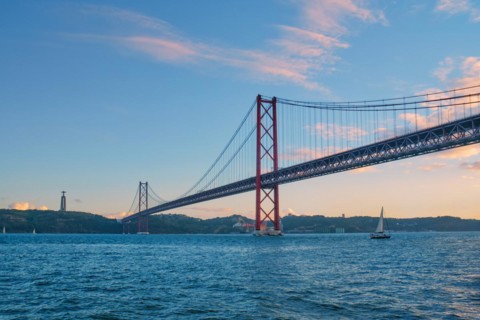View of 25 de Abril Bridge famous tourist landmark of Lisbon connecting Lisboa to Almada on Setubal Peninsula over Tagus river with tourist yacht boats passing under. Lisbon