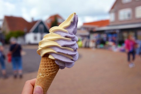 Close up of hand holding cone with mixed yellow and purple mango and berry soft serve ice cream with blurry city scene in background