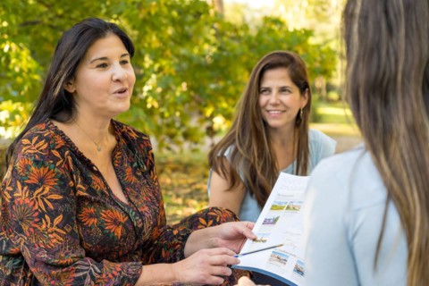 A female travel agent holds up an itinerary and explains vacation packages to two friends who listen attentively and excitedly