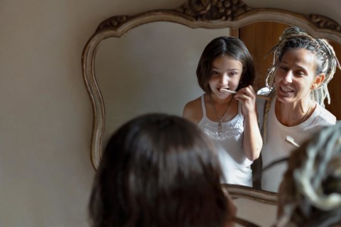 Girl looks in the mirror and brushes her teeth while her mother smiles at her. Image reflected in the bathroom mirror