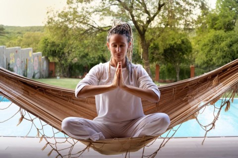Blonde yogi woman with dreadlocks in prayer position sitting in a woven hammock