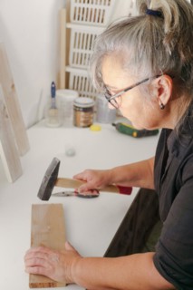 Woman seen in profile hammering a nail with a hammer into a wooden plank in her workshop
