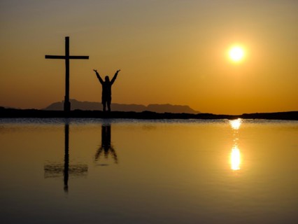 Silhouette of a woman with arms stretched up next to a cross on a summit at sunset