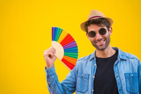 Portrait of a caucasian man in a hat smiling with a rainbow lgbt fan on a yellow background