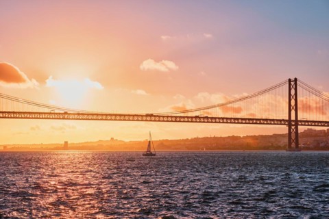 View of 25 de Abril Bridge famous tourist landmark of Lisbon connecting Lisboa and Almada on Setubal Peninsula over Tagus river with tourist yacht silhouette at sunset