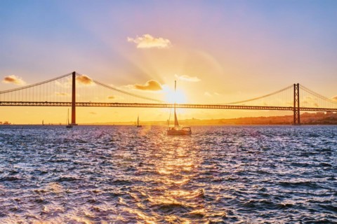 View of 25 de Abril Bridge famous tourist landmark of Lisbon connecting Lisboa and Almada on Setubal Peninsula over Tagus river with tourist yacht silhouette at sunset