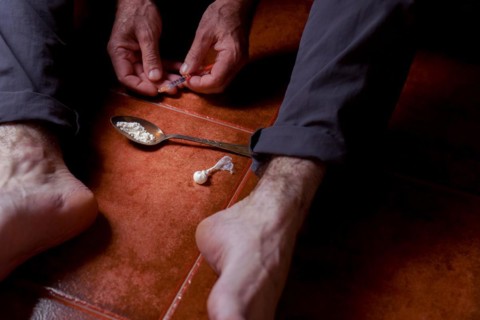 Front view of a man sitting barefoot on the floor with a heroin syringe in his hand Concept of drug addiction